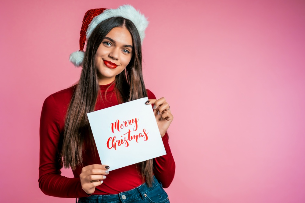 Attractive girl in Santa hat with nameplate Merry Christmas. Portrait of lady on pink background. New year concept. Happy pretty woman smiling to camera. Attractive girl in Santa hat nameplate Merry Christmas. Portrait of lady on pink