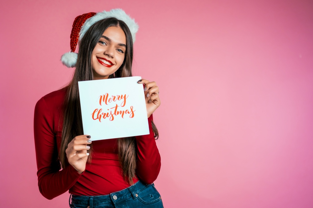 Attractive girl in Santa hat with nameplate Merry Christmas. Portrait of lady on pink background. New year concept. Happy pretty woman smiling to camera. Attractive girl in Santa hat nameplate Merry Christmas. Portrait of lady on pink