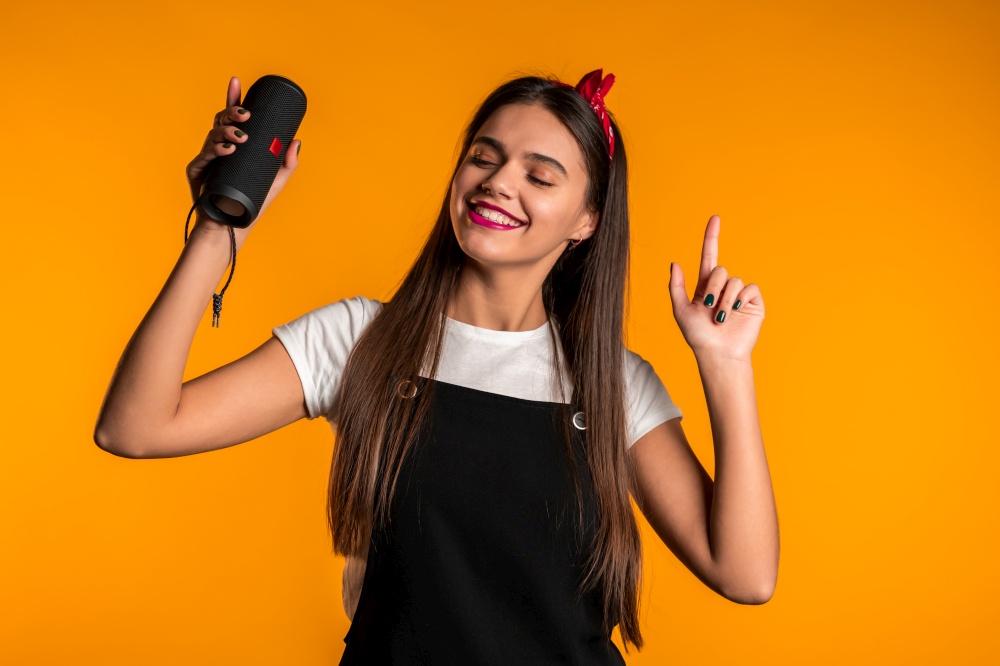 Cute woman with long hair smiling with wireless portable speaker in studio on yellow background. Music, dance concept.. Cute woman with long hair smiling with wireless portable speaker in studio on yellow background. Music, dance concept