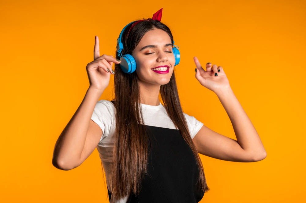 Pretty young girl with long hair having fun, smiling, dancing with headphones in studio against yellow background. Music, dance, radio concept. Pretty young girl with long hair having fun, smiling, dancing with headphones in studio against yellow background. Music, dance, radio concept,