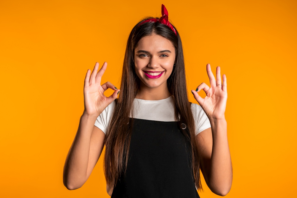 Young woman with perfect make-up making OK sign over yellow background. Winner. Success. Positive girl smiles to camera. Body language. Young woman with perfect make-up making OK sign over yellow background. Winner. Success. Positive girl smiles to camera. Body language.