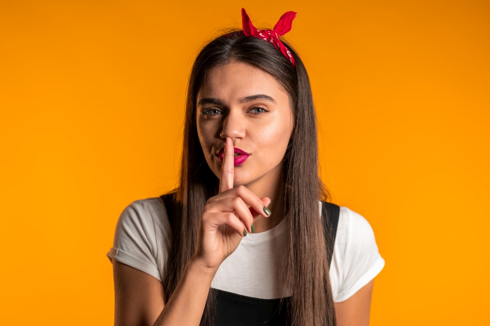 Smiling woman with long hair holding finger on her lips over yellow background. Gesture of shhh, secret, silence. Close up. Smiling woman with long hair holding finger on her lips over yellow background. Gesture of shhh, secret, silence. Close up.