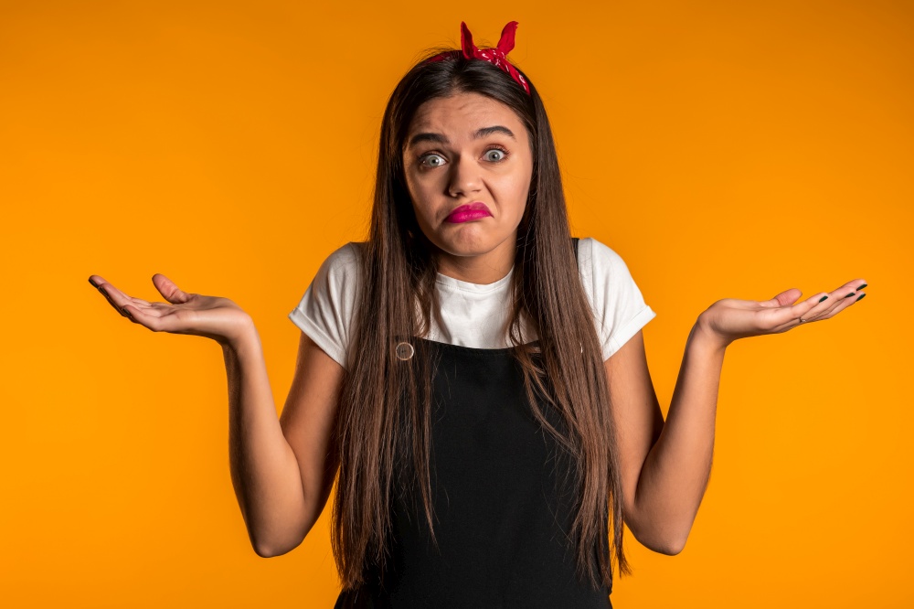 Young unsure girl shrugs her arms, makes gesture of I don&rsquo;t know, care, can&rsquo;t help anything .Young millenial woman on yellow background..  Young unsure girl shrugs her arms, makes gesture of I don&rsquo;t know, care, can&rsquo;t help anything .Young millenial woman on yellow background