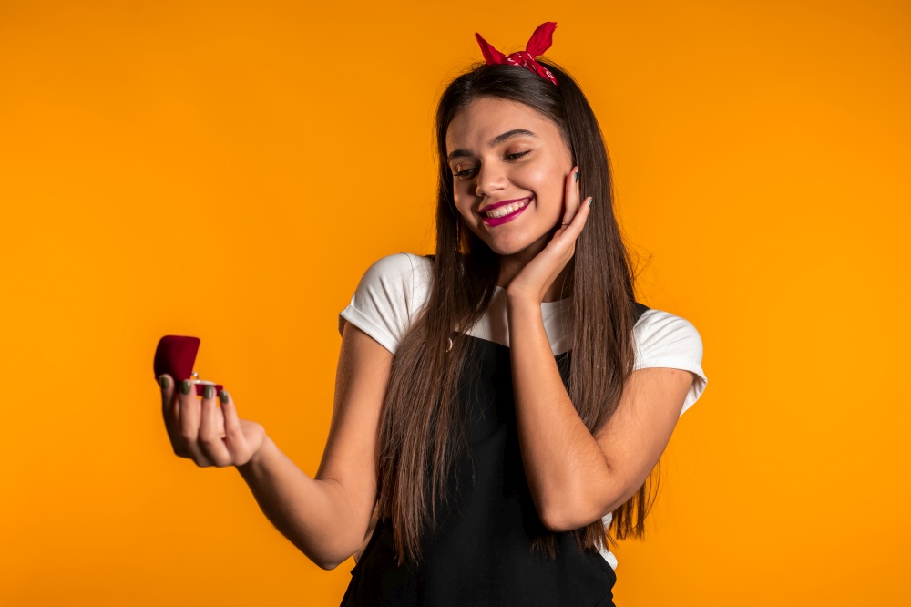 Pretty european young woman holding small jewelry box with proposal diamond ring on yellow wall background. Girl smiling, she is happy to get present, proposition for marriage. Pretty european young woman holding small jewelry box with proposal diamond ring on yellow wall background. Girl smiling, she is happy to get present, proposition for marriage.