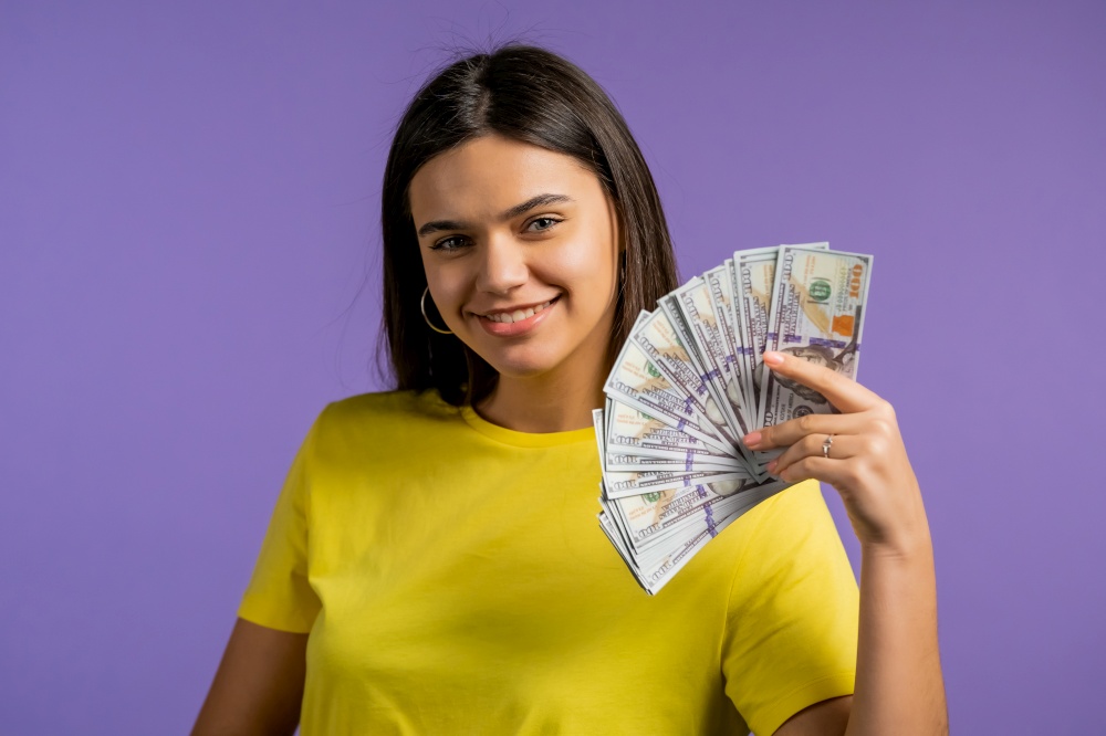 Smiling happy woman with cash money - USD currency dollars banknotes on violet wall. Symbol of jackpot, gain, victory, winning the lottery.. Smiling happy woman with cash money - USD currency dollars banknotes on violet wall. Symbol of jackpot, gain, victory, winning the lottery