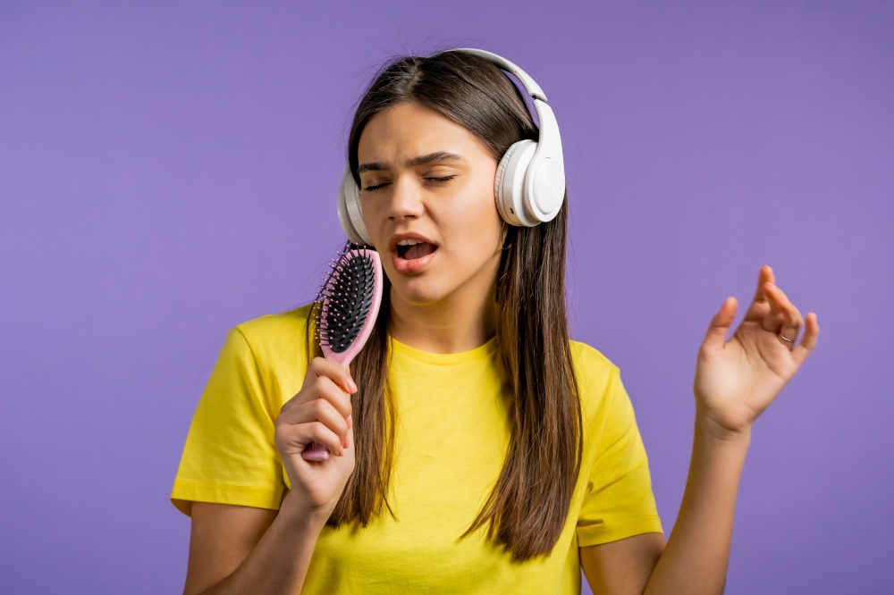 European woman singing and dancing with hair brush or comb instead microphone at purple studio background. Lady in headphones having fun, listening to music, dreams of being celebrity. High quality. European woman singing and dancing with hair brush or comb instead microphone at violet studio background. Lady in headphones having fun, listening to music, dreams of being celebrity.