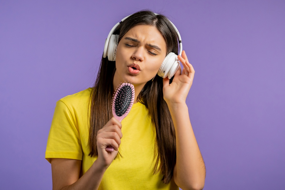 European woman singing and dancing with hair brush or comb instead microphone at purple studio background. Lady in headphones having fun, listening to music, dreams of being celebrity. High quality. European woman singing and dancing with hair brush or comb instead microphone at violet studio background. Lady in headphones having fun, listening to music, dreams of being celebrity.
