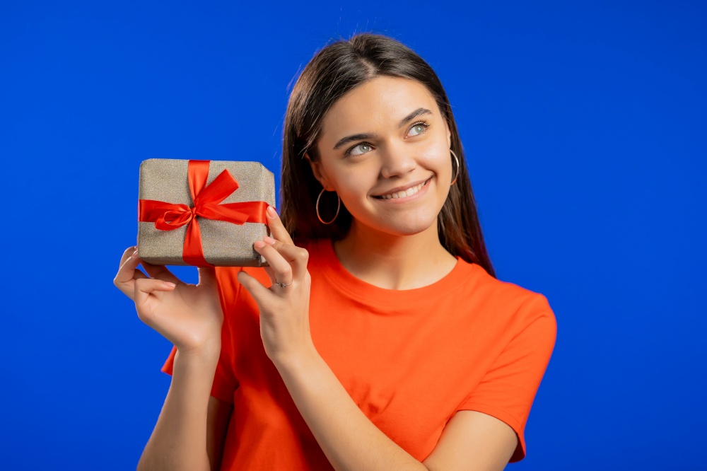 Excited woman received gift box with bow. She is happy and flattered by attention. Girl smiling with present on blue background. Studio portrait.. Excited woman received gift box with bow. She is happy and flattered by attention. Girl smiling with present on blue background. Studio portrait