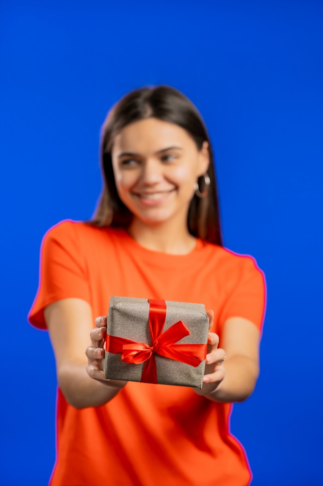 Excited woman holding gift box and gives it by hands to camera on blue wall background. Girl smiling, she is happy with present. Studio portrait.. Excited woman holding gift box and gives it by hands to camera on blue wall background. Girl smiling, she is happy with present. Studio portrait