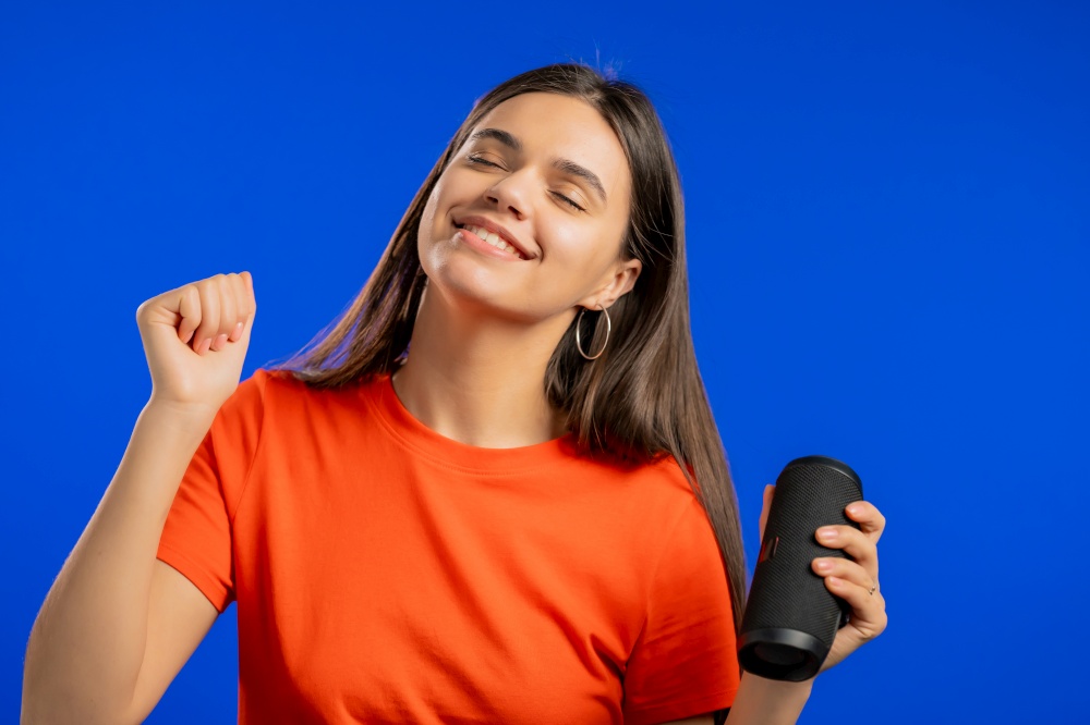 Attractive woman listening to music by wireless portable speaker - modern sound system. Lady dancing, enjoying on blue studio background. She moves to the rhythm of music. High quality. Attractive woman listening to music by wireless portable speaker - modern sound system. Lady dancing, enjoying on blue studio background. She moves to the rhythm of music.