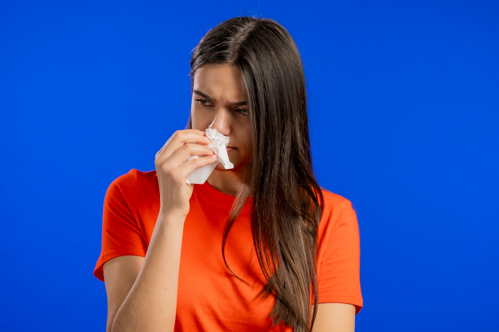 Woman sneezes into tissue. Isolated girl on blue studio background. Lady is sick, has a cold or allergic reaction. Coronavirus, epidemic 2021, illness concept.. Woman sneezes into tissue. Isolated girl on blue studio background. Lady is sick, has a cold or allergic reaction. Coronavirus, epidemic 2021, illness concept