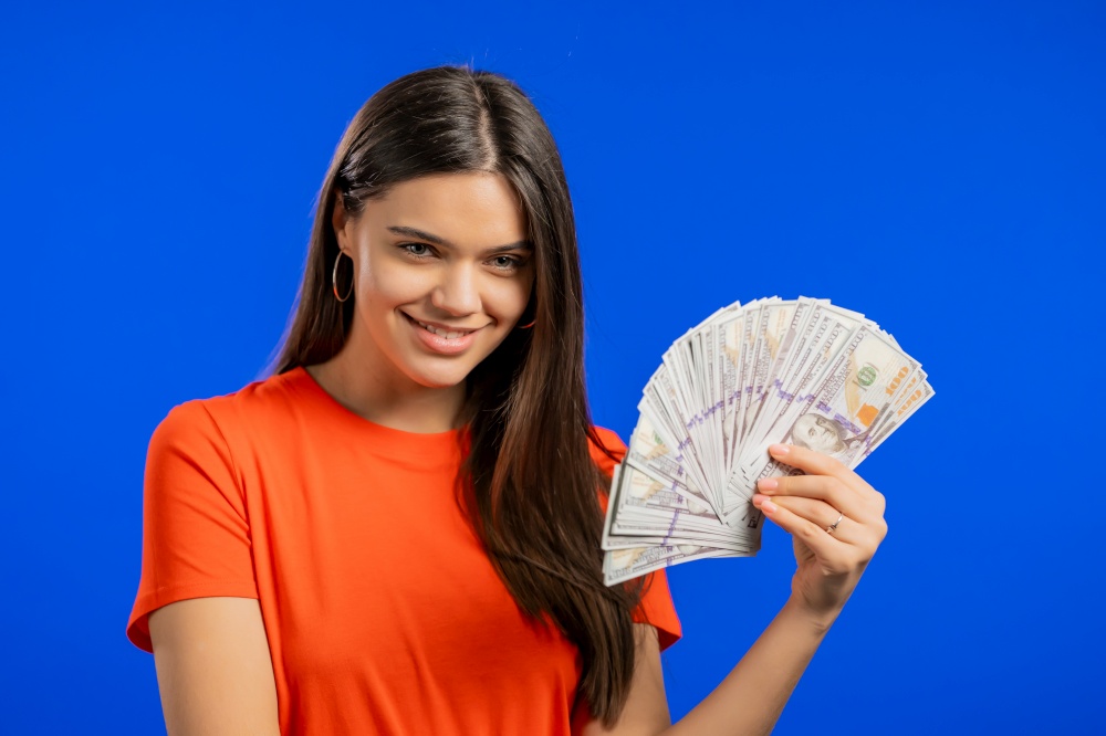 Smiling happy woman with cash money - USD currency dollars banknotes on blue wall. Symbol of jackpot, gain, victory, winning the lottery.. Smiling happy woman with cash money - USD currency dollars banknotes on blue wall. Symbol of jackpot, gain, victory, winning the lottery