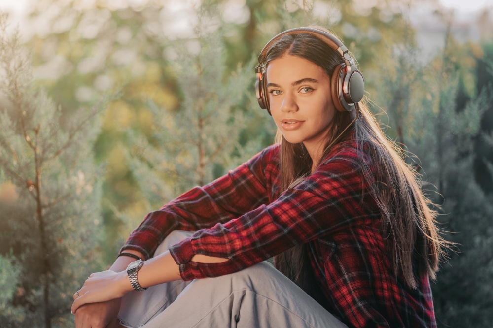 Young teenager listens to music through headphones in park.Girl in red plaid shirt smiles, dancing to rhythm.Concept of student life, freedom, modern youth.. Young teenager listens to music through headphones in park.Girl in red plaid shirt smiles, dancing to rhythm.Concept of student life, freedom, modern youth