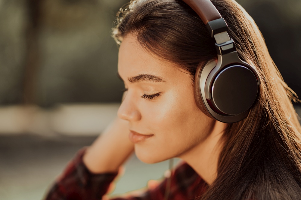 Cute girl&rsquo;s face with headphones. Young woman enjoys music while sitting in green park. Concept of youth, freedom, technology.. Cute girl&rsquo;s face with headphones. Young woman enjoys music while sitting in green park. Concept of youth, freedom, technology