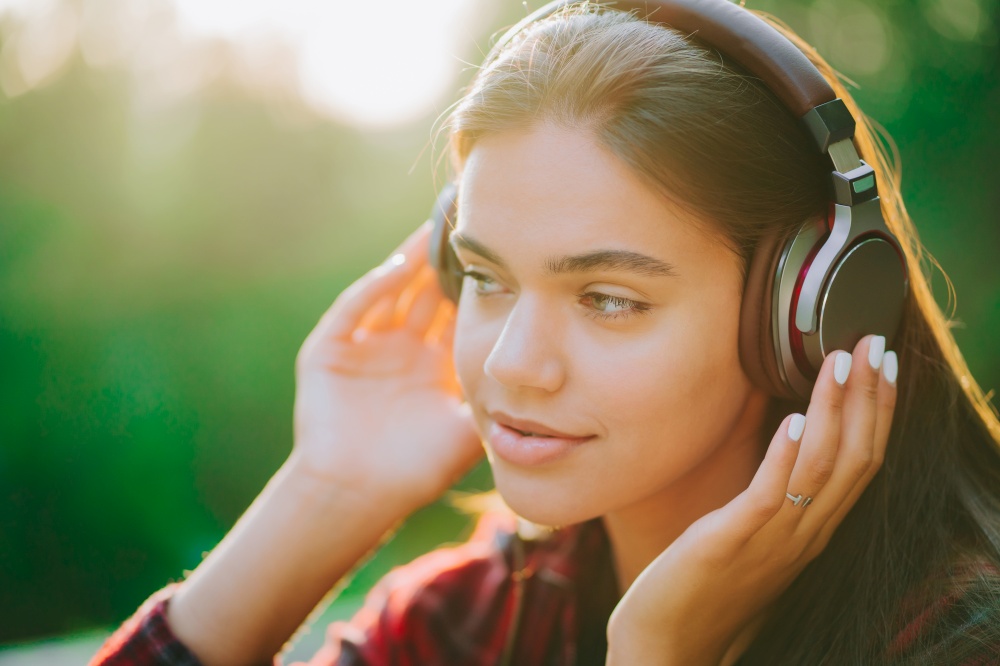 Cute girl&rsquo;s face with headphones. Young woman enjoys music while sitting in green park. Concept of youth, freedom, technology.. Cute girl&rsquo;s face with headphones. Young woman enjoys music while sitting in green park. Concept of youth, freedom, technology