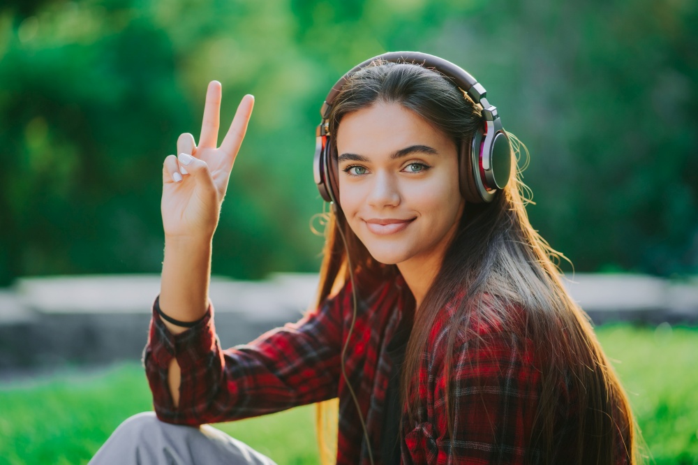 Young teenager listens to music through headphones in park.Girl in red plaid shirt smiles, dancing to rhythm.Concept of student life, freedom, modern youth.. Young teenager listens to music through headphones in park.Girl in red plaid shirt smiles, dancing to rhythm.Concept of student life, freedom, modern youth