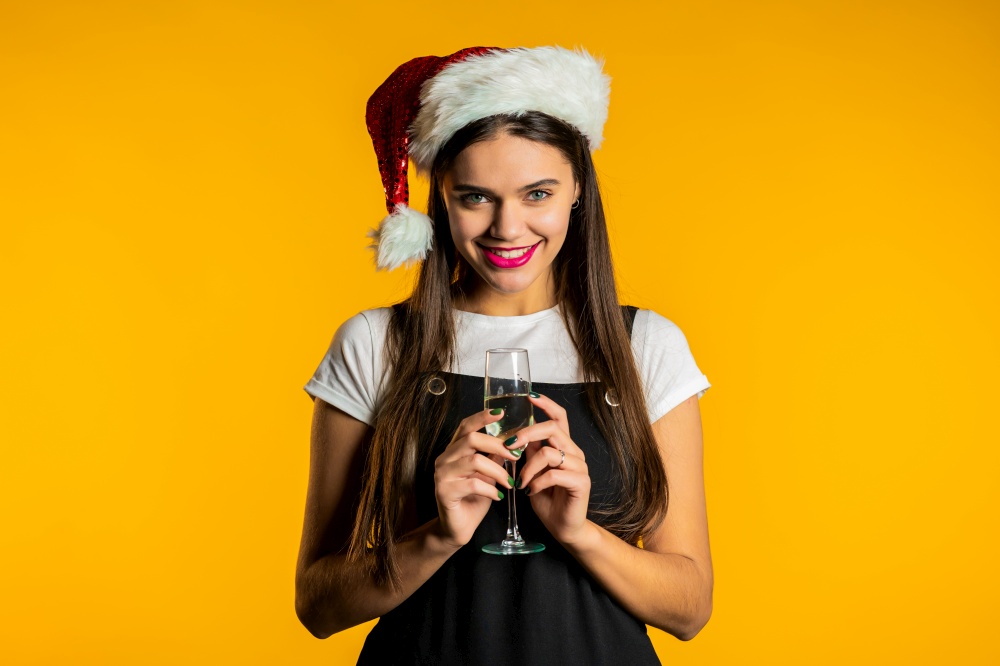 Young woman smiling and holding glass of champagne or wine on yellow studio background. Lady in Santa hat. Christmas mood. High quality photo. Young woman smiling and holding glass of champagne or wine on yellow studio background. Lady in Santa hat. New year mood.