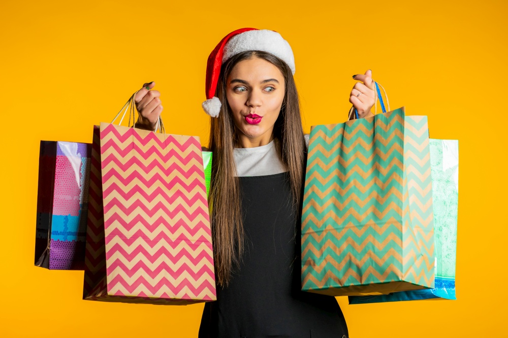 Happy woman in Santa hat with colorful paper bags after shopping on yellow studio background. Winter sale, purchases, spending money on gifts concept. High quality photo. Happy woman in Santa hat with colorful paper bags after shopping on yellow studio background. Winter sale, purchases, spending money on gifts concept.