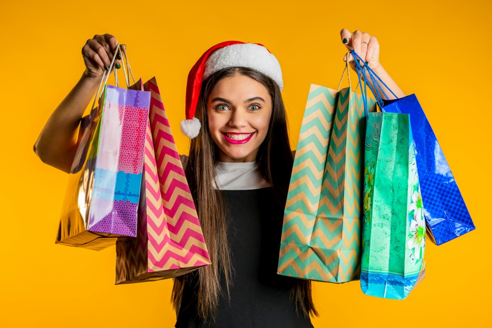 Happy woman in Santa hat with colorful paper bags after shopping on yellow studio background. Winter sale, purchases, spending money on gifts concept. High quality photo. Happy woman in Santa hat with colorful paper bags after shopping on yellow studio background. Winter sale, purchases, spending money on gifts concept.