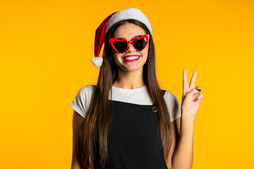 Beautiful woman with long hair in Santa hat showing friendship gesture, hand in peace sign. V - victory icon on studio yellow background. High quality photo. Beautiful woman with long hair in Santa hat showing friendship gesture, hand in peace sign. V - victory icon on studio yellow background.