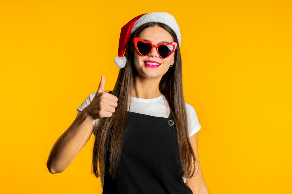 Positive woman in Santa hat with like hand sign, thumb up gesture. Happy girl, correct perfect choice, great deal, yellow background. Positive lady smiles to camera, approval. High quality photo. Positive woman in Santa hat with like hand sign, thumb up gesture. Happy girl, correct perfect choice, great deal, yellow background. Positive lady smiles to camera, approval, trust concept