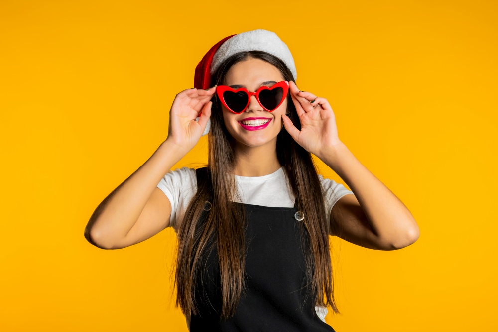 Beautiful woman with long hair in Santa hat and red heart-shaped sunglasses on studio yellow background. High quality photo. Beautiful woman with long hair in Santa hat and red heart-shaped sunglasses on studio yellow background.