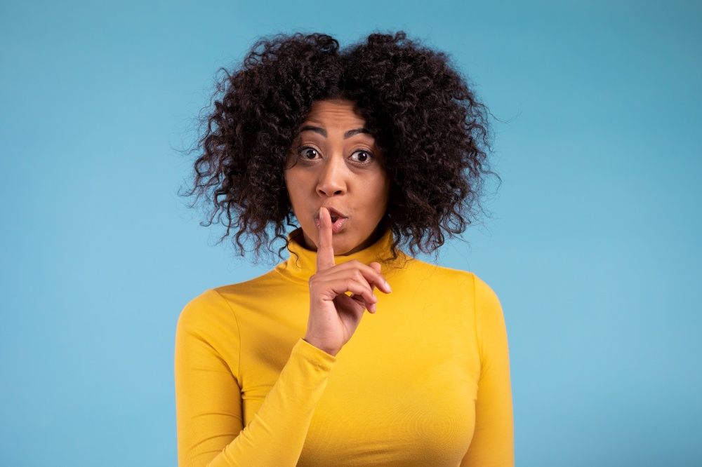 Smiling african woman with curly hair holding finger on her lips over blue background. Gesture of shhh, secret, silence. Close up.. Smiling african woman with curly hair holding finger on her lips over blue background. Gesture of shhh, secret, silence. Close up