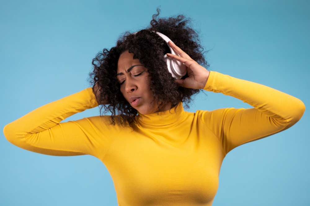 African woman dancing with wireless headphones isolated on blue studio wall. Cute lady in yellow wear, she is singing. Music, radio, happiness, freedom, youth concept. African woman dancing with wireless headphones isolated on blue studio wall. Cute lady in yellow wear, she is singing. Music, radio, happiness, freedom, youth concept.