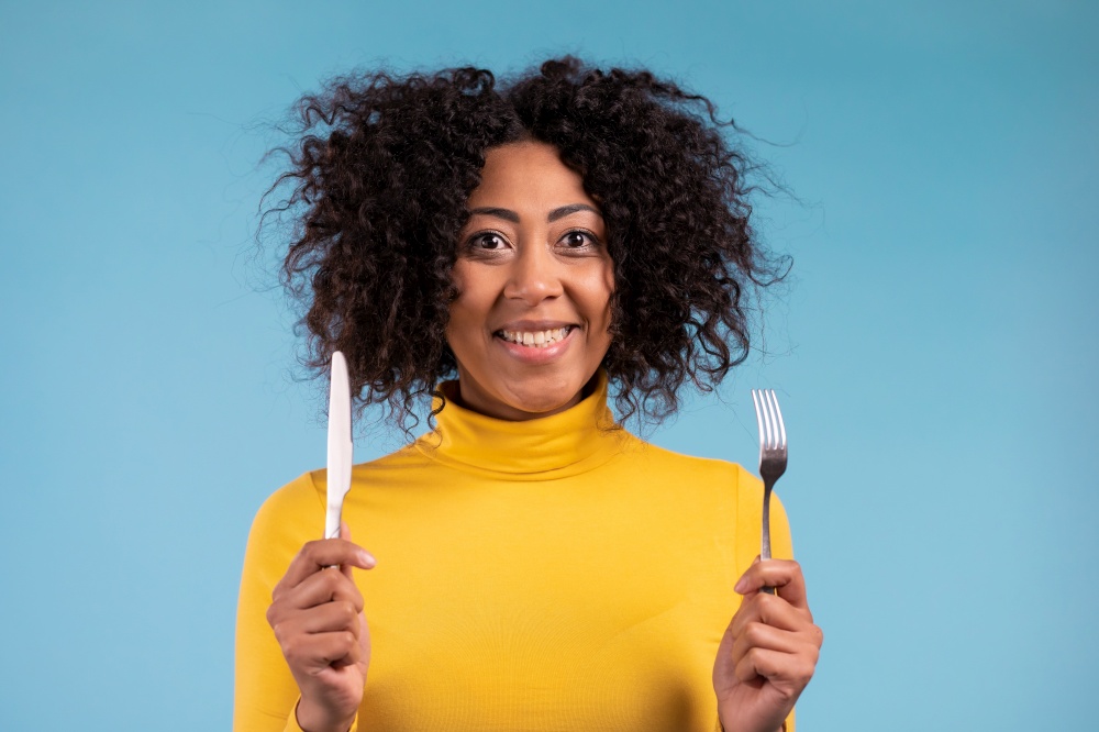Portrait of hungry african woman with fork and knife. Lady waiting for serving dinner dishes with cutlery on blue studio background. High quality photo. Portrait of hungry african woman with fork and knife. Lady waiting for serving dinner dishes with cutlery on blue studio background.