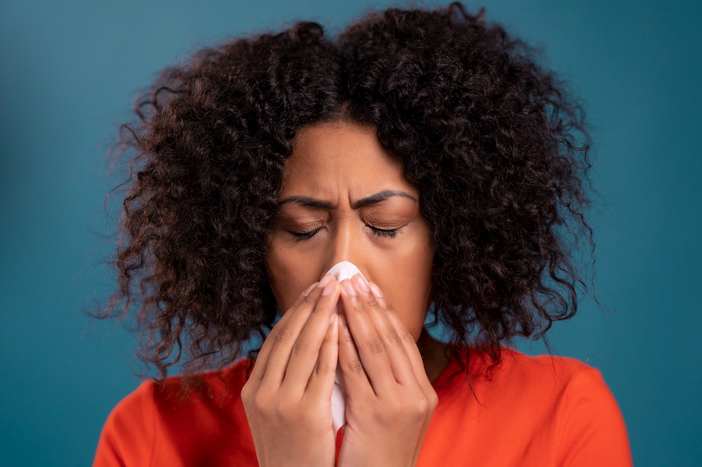 African woman sneezes into tissue. Isolated girl on blue studio background. Lady is sick, has a cold or allergic reaction. Coronavirus, epidemic 2021, illness concept.. African woman sneezes into tissue. Isolated girl on blue studio background. Lady is sick, has a cold or allergic reaction. Coronavirus, epidemic 2021, illness concept