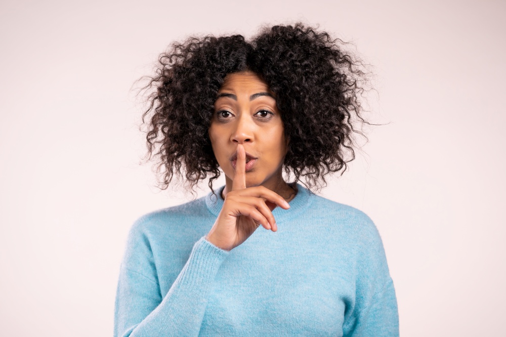 Smiling african woman with curly hair holding finger on her lips over white background. Gesture of shhh, secret, silence. Close up. Smiling african woman with curly hair holding finger on her lips over white background. Gesture of shhh, secret, silence. Close up.