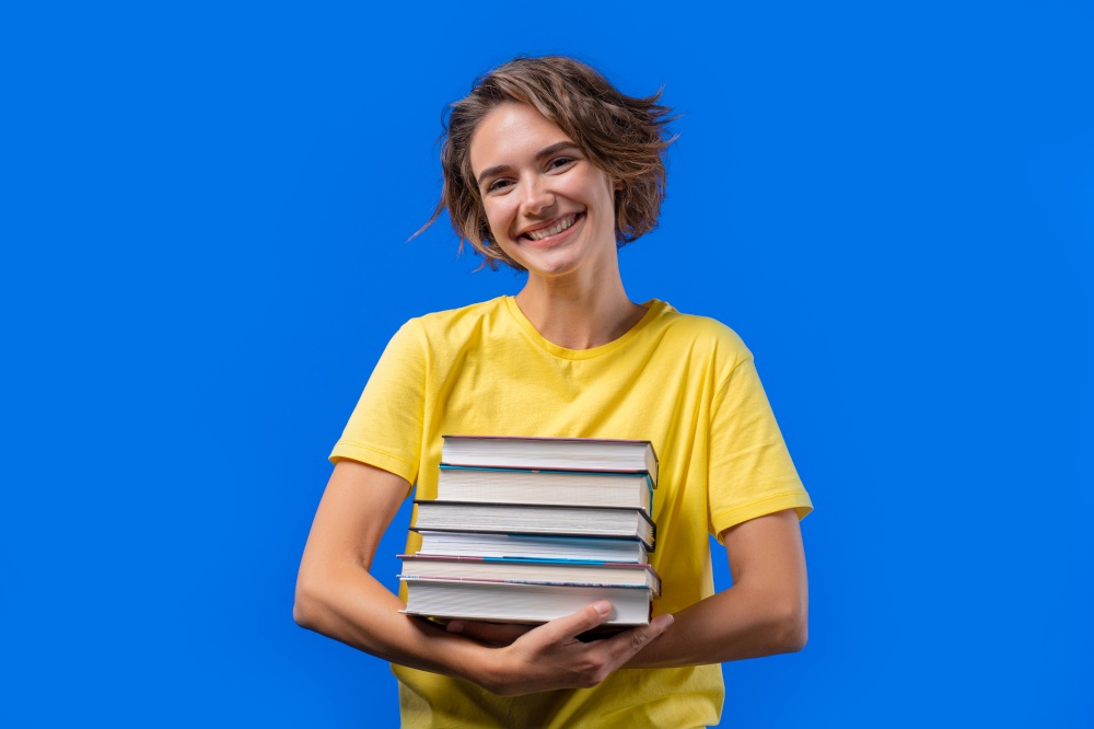 Clever student woman holds stack of university books from college library on blue background. Happy girl smiles, she is happy to graduate. High quality photo. Clever student woman holds stack of university books from college library on blue background. Happy girl smiles, she is happy to graduate.