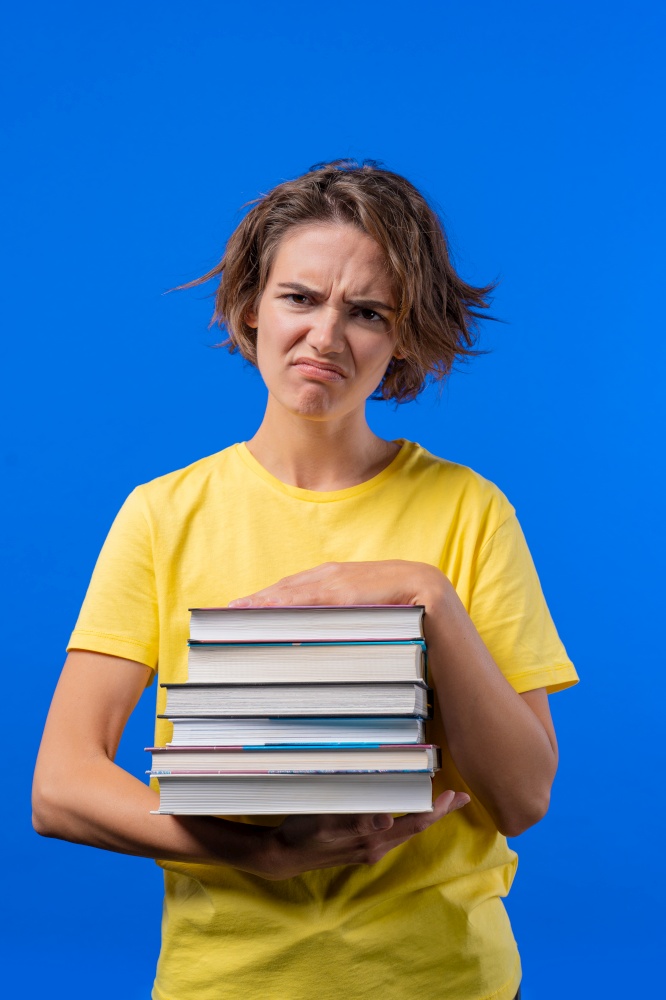 Lazy woman student is dissatisfied with amount of books homework on blue background. Girl with short hair in displeasure, she is annoyed, discouraged frustrated by studies. High quality photo. Lazy woman student is dissatisfied with amount of books homework on blue background. Girl with short hair in displeasure, she is annoyed, discouraged frustrated by studies