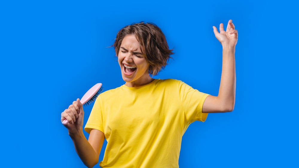 Woman singing, dancing with hair brush instead microphone on blue background. Lady having fun, listening to music, karaoke, dreams of being celebrity. High quality photo. Woman singing, dancing with hair brush instead microphone on blue background. Lady having fun, listening to music, karaoke, dreams of being celebrity.