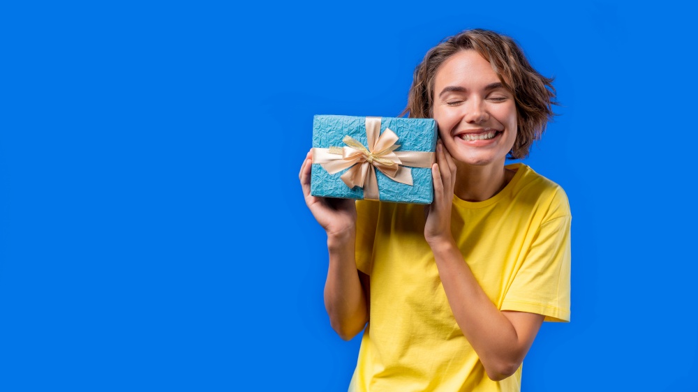 Happy birthday woman with gift box on blue background. Girl smiling, she is happy with present. Copy space. High quality photo. Happy birthday woman with gift box on blue background. Girl smiling, she is happy with present. Copy space