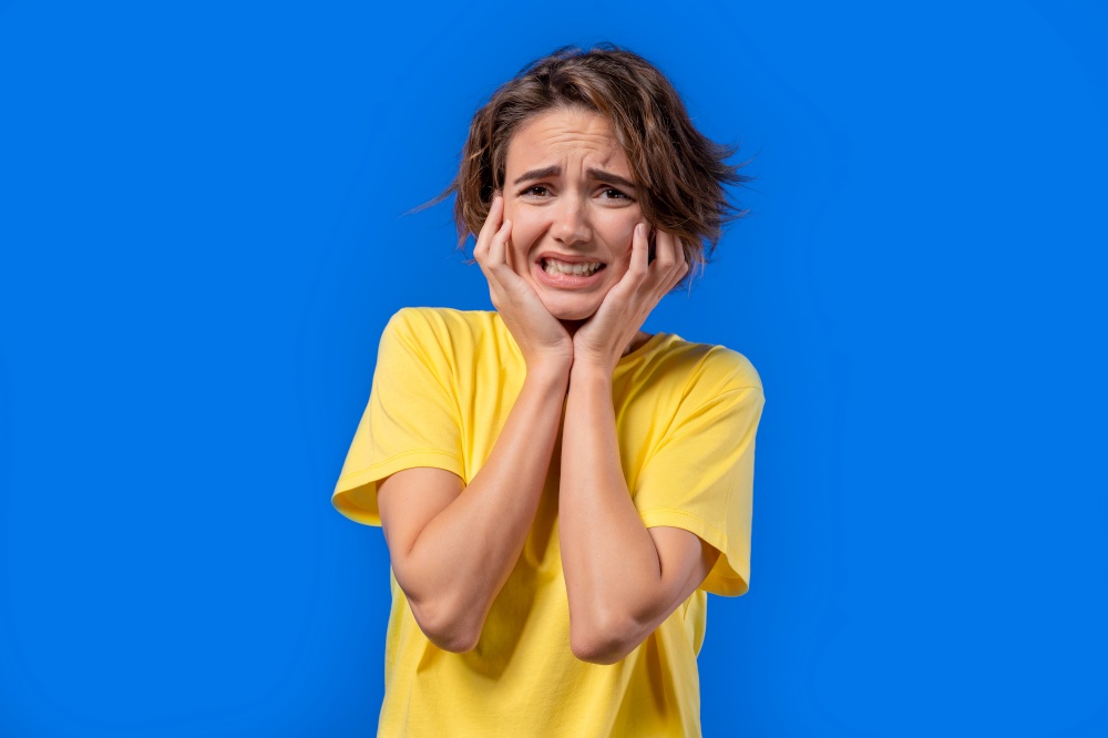 Woman afraid of something and looks into camera with big eyes full of pain on blue background. Phobia, trouble, panic concept. High quality photo. Woman afraid of something and looks into camera with big eyes full of pain on blue background. Phobia, trouble, panic concept.