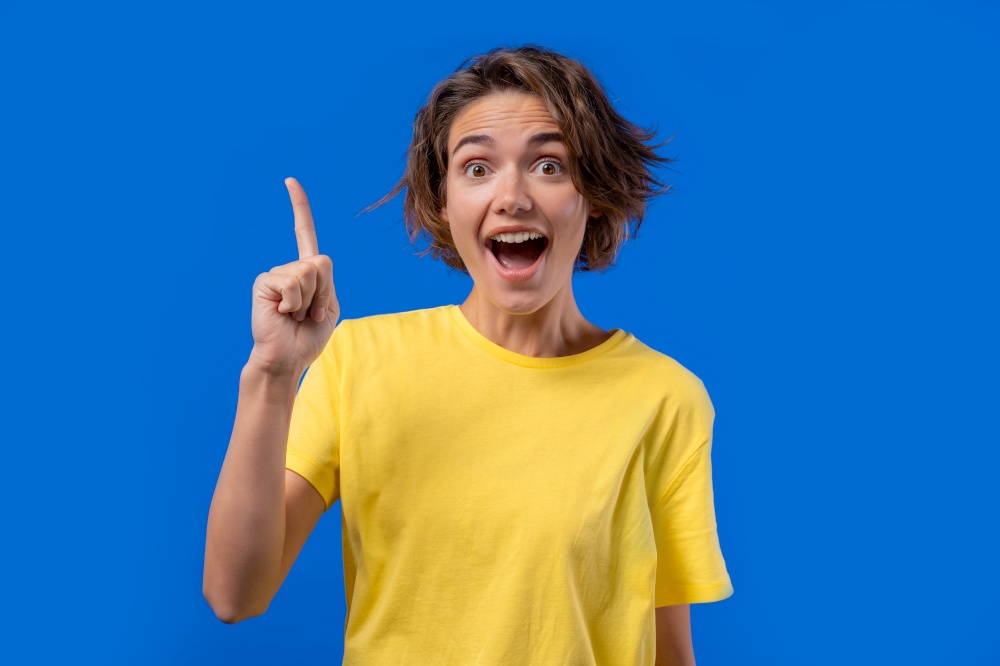Happy woman having idea eureka moment, pointing finger up on blue background. Smart student girl showing answer gesture or remembered what she forgot, memory concept. High quality photo. Happy woman having idea eureka moment, pointing finger up on blue background. Smart student girl showing answer gesture or remembered what she forgot, memory concept