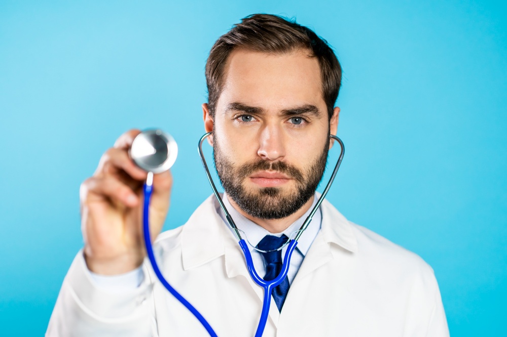 Portrait of young handsome man in professional medical white coat using stethoscope isolated on blue studio background. Man points his instrument at camera as if he is listening to ill patient.. Portrait of young handsome man in professional medical white coat using stethoscope isolated on blue studio background. Man points his instrument at camera as if he is listening to ill patient