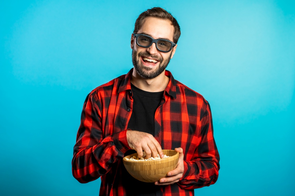 Young european manl in 3d glasses watching fascinating comedy movie, laughing and eating popcorn on blue studio background. Young european manl in 3d glasses watching fascinating comedy movie, laughing and eating popcorn on blue studio background.