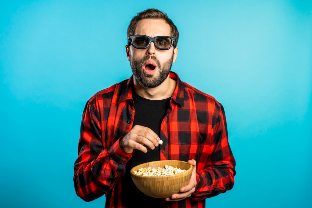 Young man in 3d glasses watching fascinating horror movie and eating popcorn on blue studio background. Young man in 3d glasses watching fascinating horror movie and eating popcorn on blue studio background.