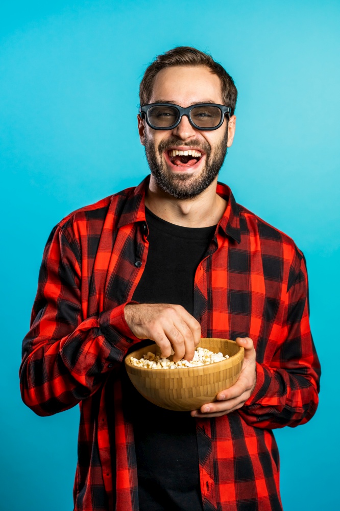 Young european manl in 3d glasses watching fascinating comedy movie, laughing and eating popcorn on blue studio background. Young european manl in 3d glasses watching fascinating comedy movie, laughing and eating popcorn on blue studio background.