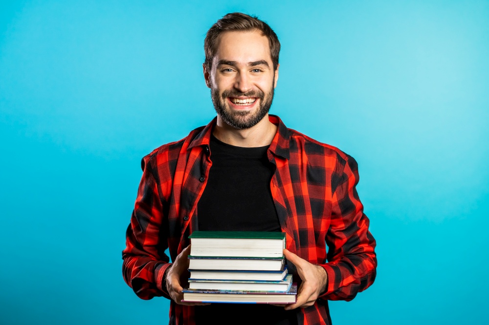 European student in red plaid shirt on blue background in studio holds stack of university books from library. Guy smiles, he is happy to graduate. European student in red plaid shirt on blue background in studio holds stack of university books from library. Guy smiles, he is happy to graduate.