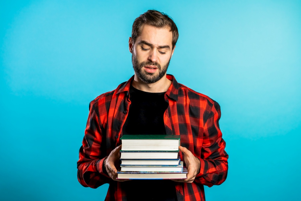 European student on blue background is dissatisfied with amount of homework and books. Man confused, he is annoyed, discouraged frustrated by studies. European student on blue background is dissatisfied with amount of homework and books. Man confused, he is annoyed, discouraged frustrated by studies.