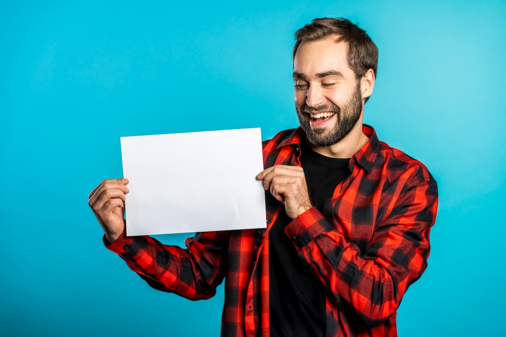 Handsome man holding horizontal white a4 paper poster. Copy space. Smiling hipster guy in red plaid shirt on blue background. Handsome man holding horizontal white a4 paper poster. Copy space. Smiling hipster guy in red plaid shirt on blue background.