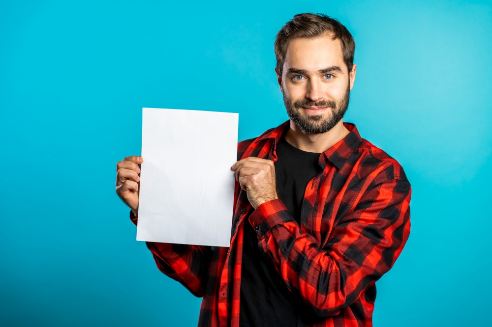 Handsome man holding vertical white a4 paper poster. Copy space. Smiling hipster guy in red plaid shirt on blue background. Handsome man holding vertical white a4 paper poster. Copy space. Smiling hipster guy in red plaid shirt on blue background.