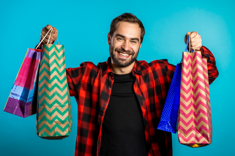 Handsome young man holds shopping paper bags on blue studio background. Guy bought presents on sales with discounts. Handsome young man holds shopping paper bags on blue studio background. Guy bought presents on sales with discounts.