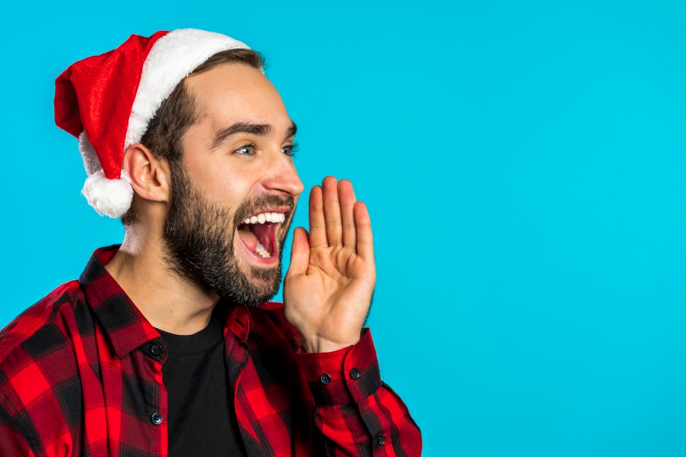 Handsome man in Santa hat on blue studio background shouting, announcing something, keeping hand on cheek, copy space. Christmas, New year concept. High quality photo. Handsome man in Santa hat on blue studio background shouting, announcing something, keeping hand on cheek, copy space. Christmas, New year concept.
