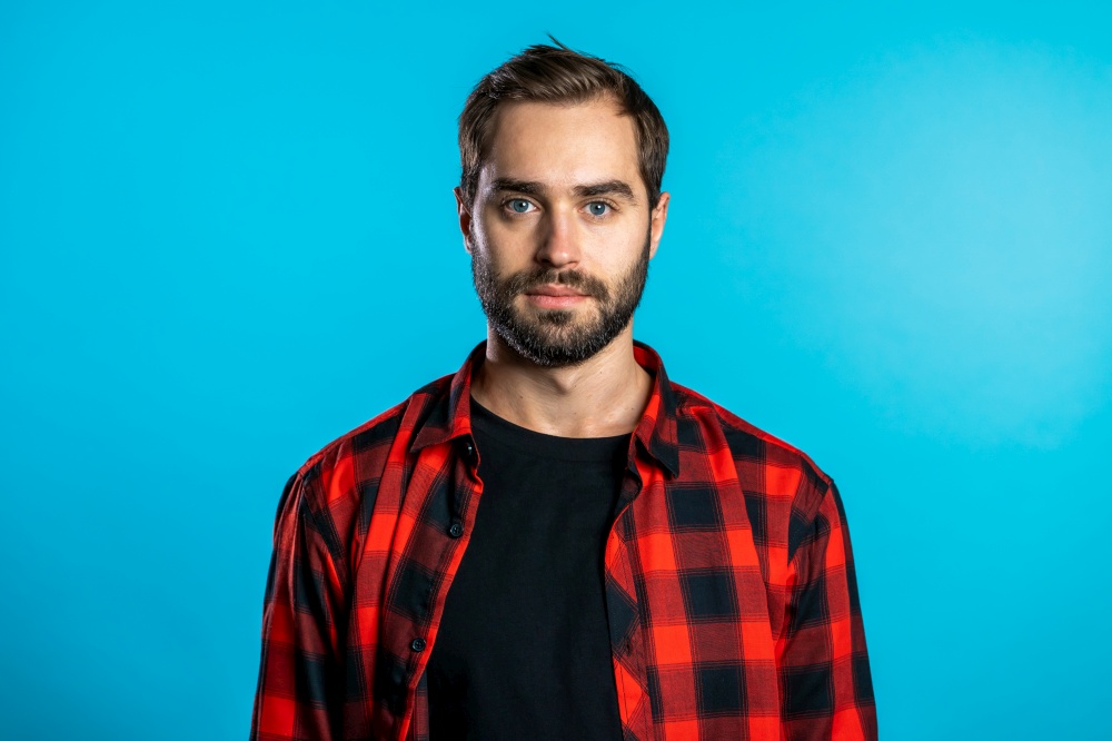Handsome european man with trendy beard in red plaid shirt on blue studio background. Cheerful guy smiling and looking to camera. Handsome serious european man with trendy beard in red plaid shirt on blue studio background. Cheerful guy smiling and looking to camera.