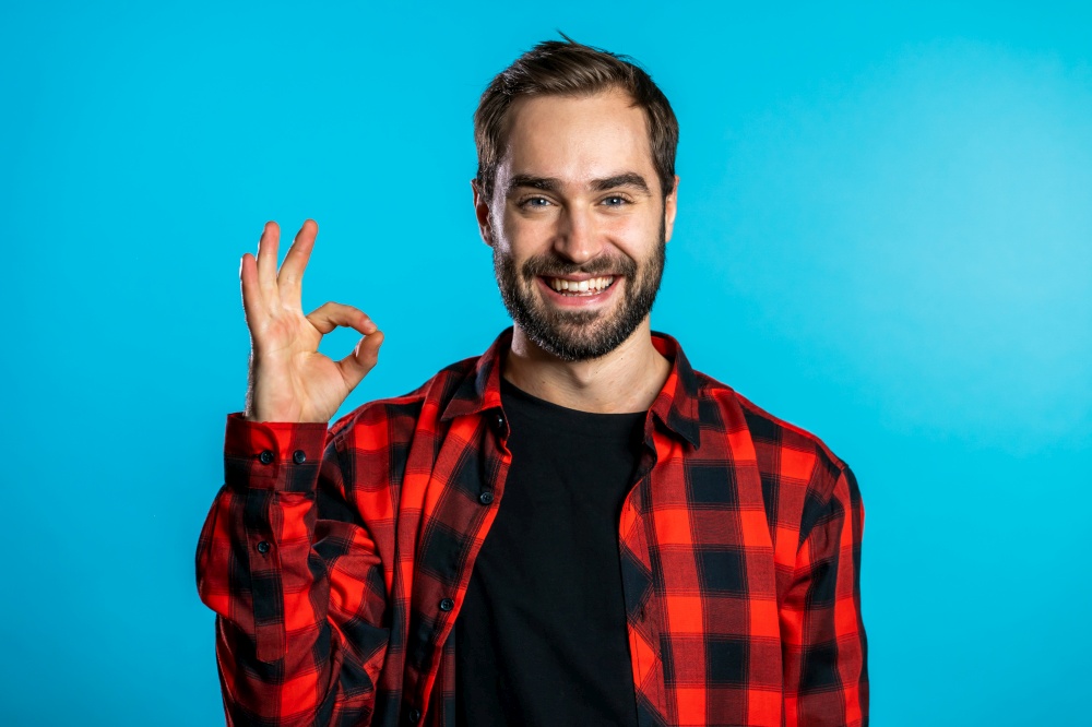 Young handsome man with beard making OK sign over blue background and smiles to camera. Young handsome man with beard making OK sign over blue background and smiles to camera.