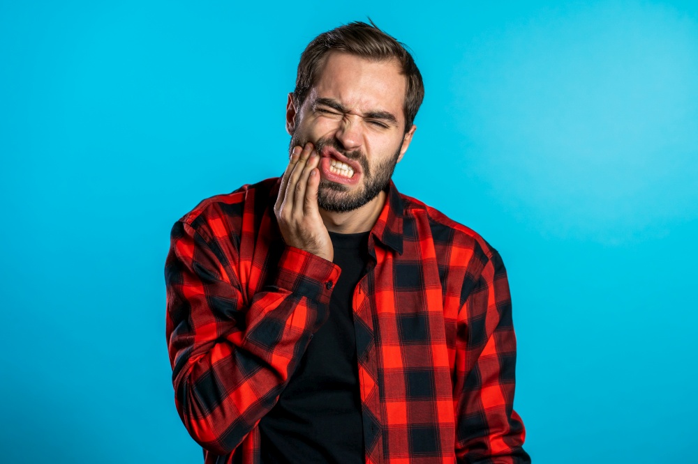 Young handsome man in red shirt with tooth pain on blue studio background. Toothache, dental problems, stomatology and medicine concept. Young handsome man in red shirt with tooth pain on blue studio background. Toothache, dental problems, stomatology and medicine concept.